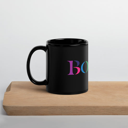 A glossy black ceramic mug resting on a light wooden surface. The mug features the word “BOSTON” in large serif letters, each filled with different bright colors including green, orange, purple, pink, and teal.