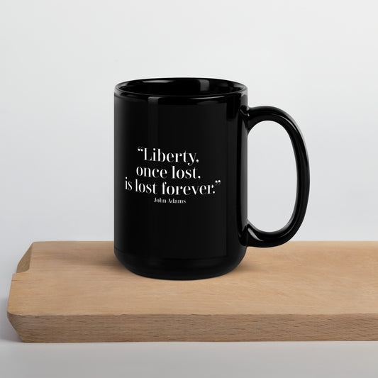 A glossy black ceramic mug featuring the quote “Liberty, once lost, is lost forever.” — John Adams, printed in white serif font on the front. The mug rests on a light wooden surface with a neutral background.