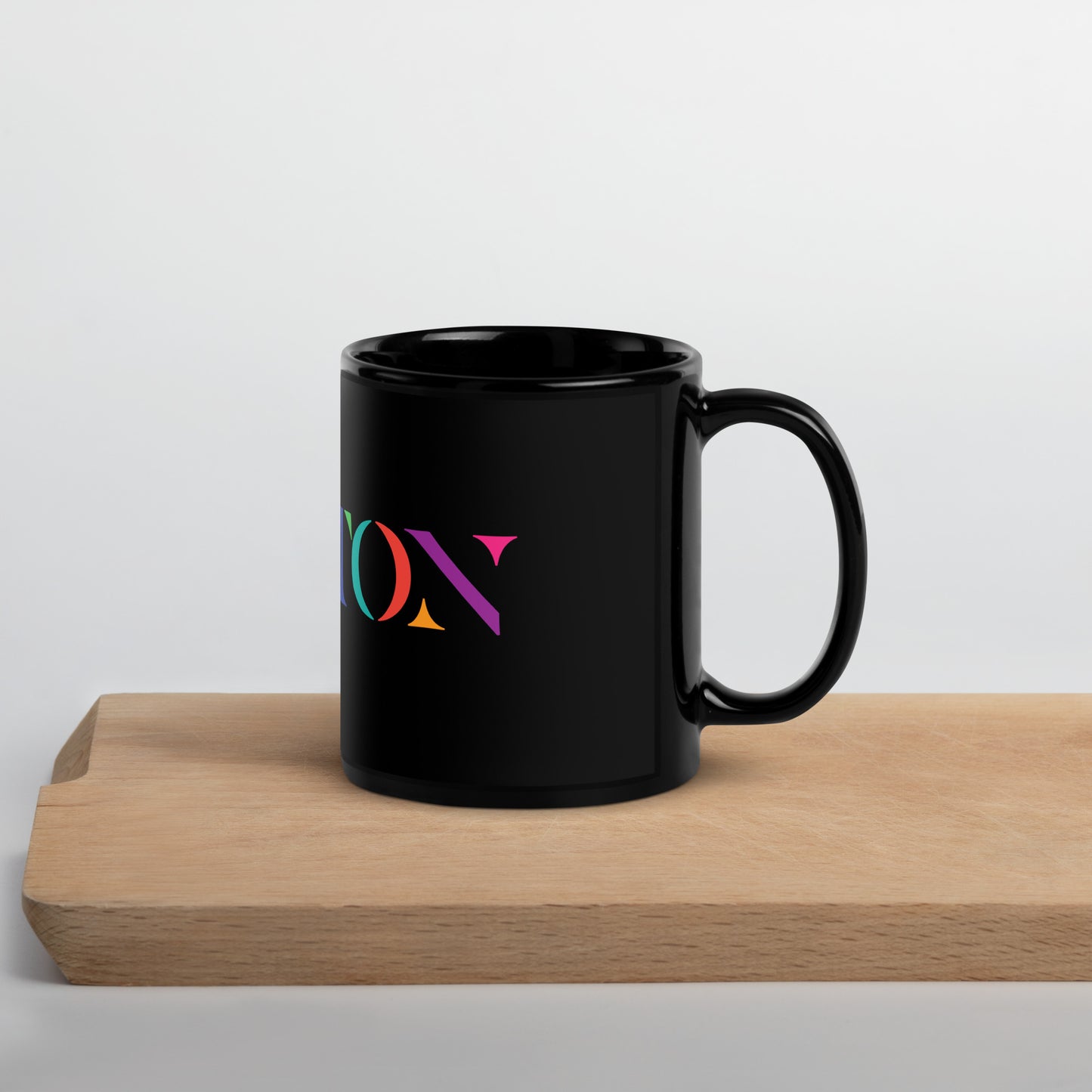 A glossy black ceramic mug resting on a light wooden surface. The mug features the word “BOSTON” in large serif letters, each filled with different bright colors including green, orange, purple, pink, and teal.