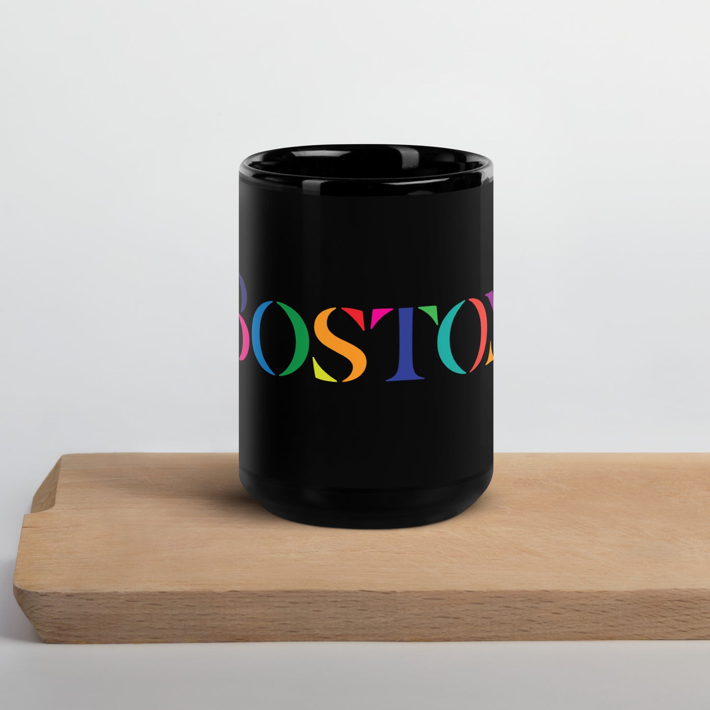 A glossy black ceramic mug resting on a light wooden surface. The mug features the word “BOSTON” in large serif letters, each filled with different bright colors including green, orange, purple, pink, and teal.