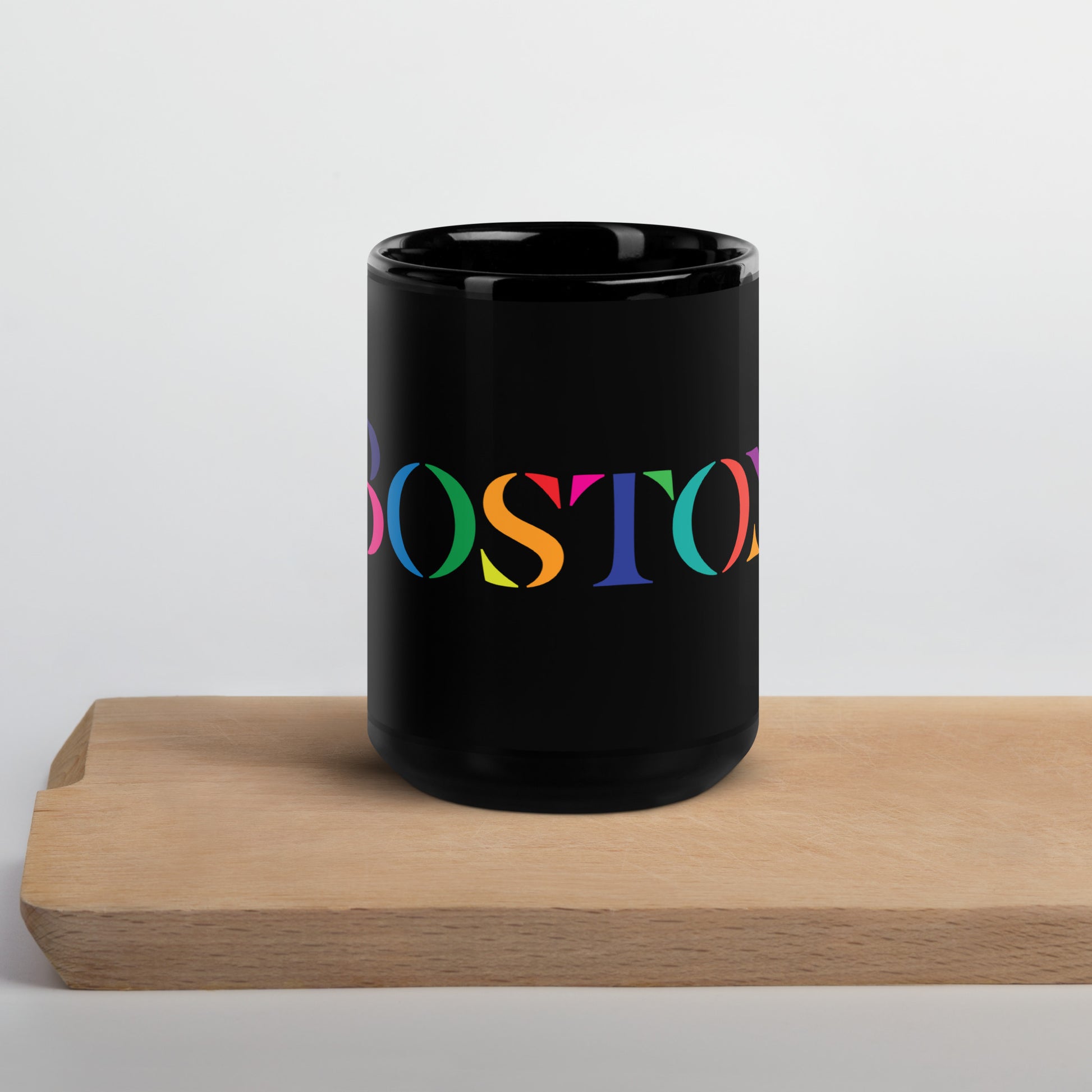 A glossy black ceramic mug resting on a light wooden surface. The mug features the word “BOSTON” in large serif letters, each filled with different bright colors including green, orange, purple, pink, and teal.