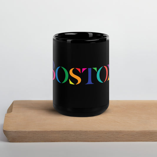 A glossy black ceramic mug resting on a light wooden surface. The mug features the word “BOSTON” in large serif letters, each filled with different bright colors including green, orange, purple, pink, and teal.