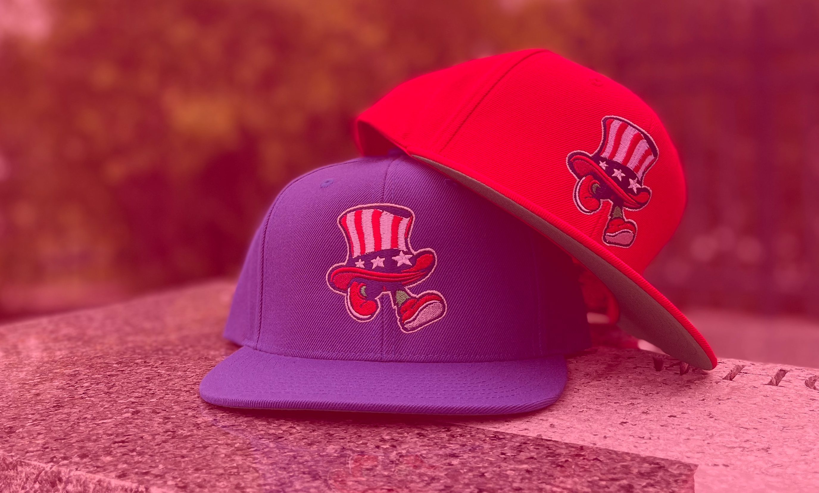 Two Snapbacks caps, one red and one Royal Blue, with a mascot logo on a granite base and blurred background.