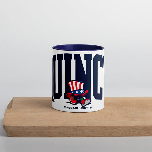 A white ceramic mug with a navy blue interior and handle, featuring bold navy letters spelling “QUINCY” across the front. In front of the text is a cartoon-style Walking President character wearing a red, white, and blue top hat and red shoes. The word “Massachusetts” appears below the artwork. The mug sits on a light wooden surface with a soft gray background.