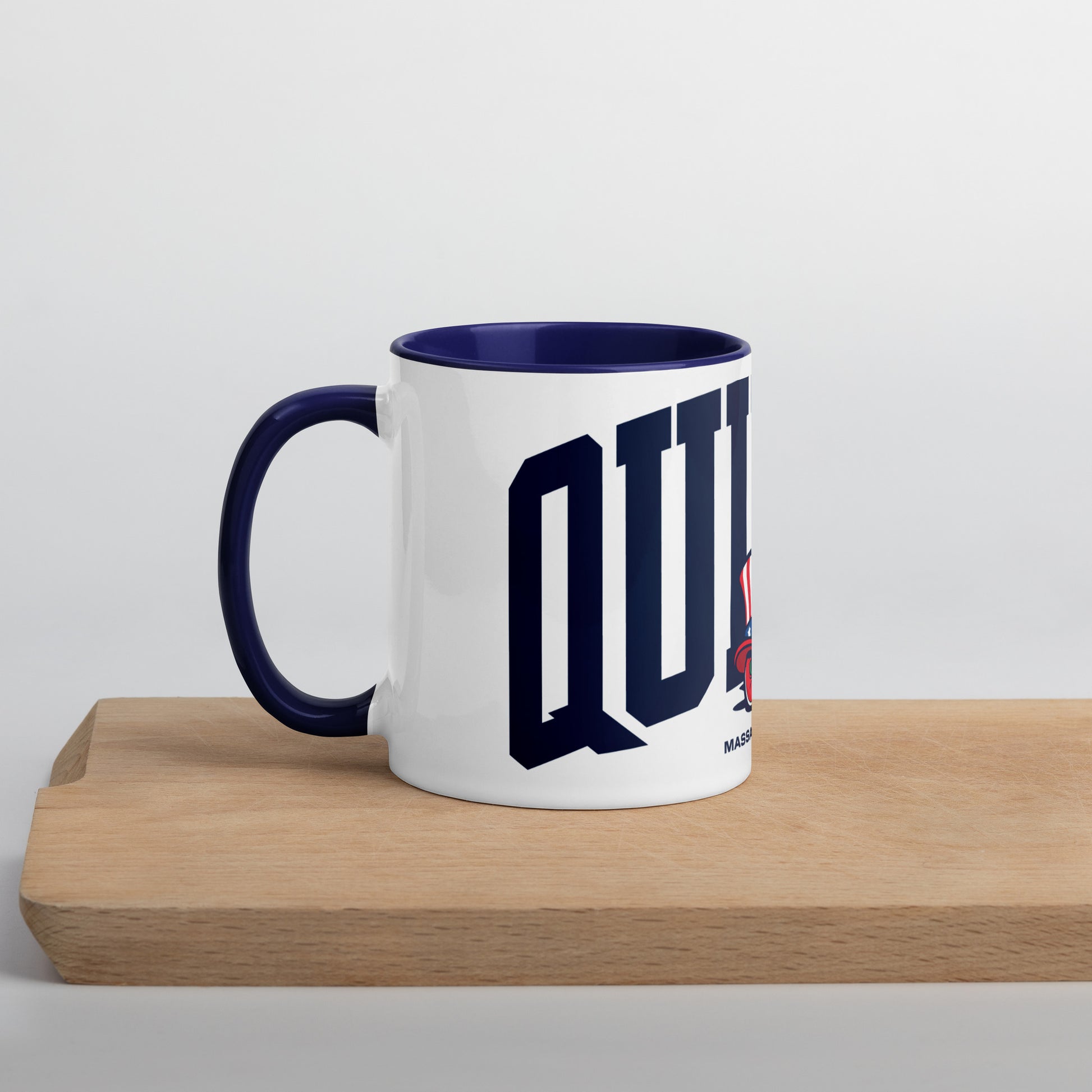 A white ceramic mug with a navy blue interior and handle, featuring bold navy letters spelling “QUINCY” across the front. In front of the text is a cartoon-style Walking President character wearing a red, white, and blue top hat and red shoes. The word “Massachusetts” appears below the artwork. The mug sits on a light wooden surface with a soft gray background.