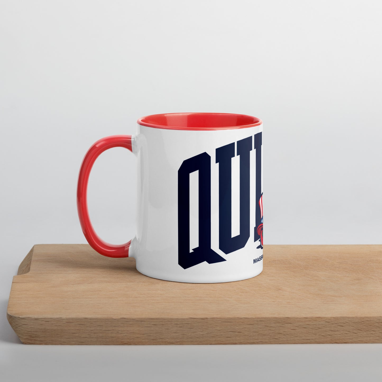 A white ceramic mug with a red interior and handle, featuring bold navy letters spelling “QUINCY” across the front. In front of the text is a cartoon-style Walking President character wearing a red, white, and blue top hat and red shoes. The word “Massachusetts” appears below the artwork. The mug sits on a light wooden surface with a soft gray background.